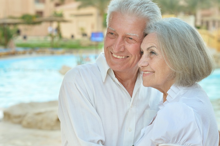 A smiling senior couple in casual clothing enjoying a peaceful outdoor moment by the water, representing companionship, love, and healthy aging.