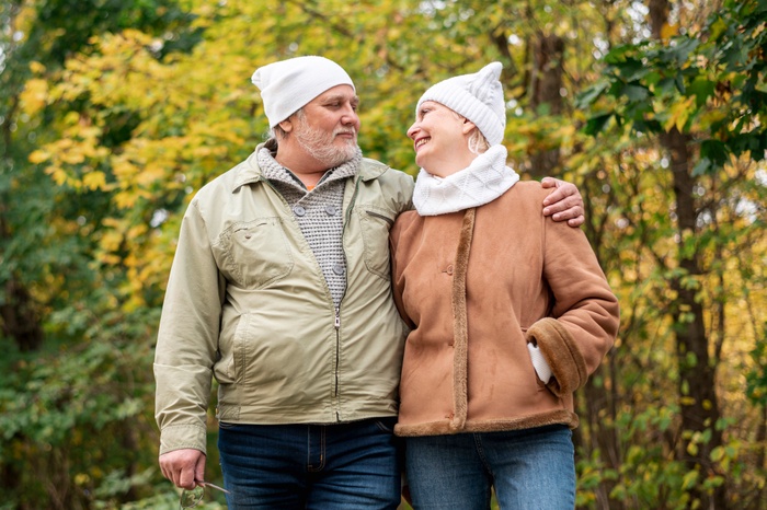 Older couple in knit hats and warm coats smiling at each other as they stand close in an autumn woodland